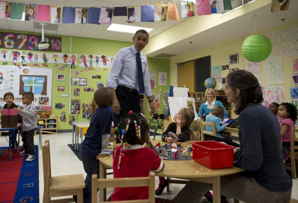 A man in a shirt and tie visiting a busy preschool classroom where children play at a table