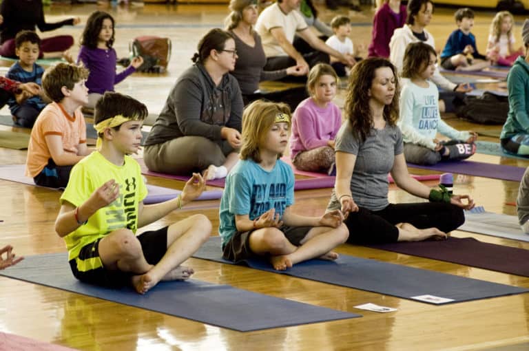 Children and adults seated cross-legged in a kids yoga class, practicing mudra-based poses and mindfulness techniques.